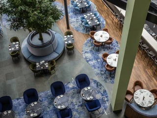 Bar Luftaufnahme einer modernen Hotelbar, ausgestattet mit runden Tischen und blauen Teppichen. Ein großer Baum im Zentrum sorgt für Atmosphäre, während bunte Stühle harmonisch arrangiert sind.Aerial view of a modern hotel bar, furnished with round tables and blue carpets. A large tree in the center creates atmosphere, while colorful chairs are harmoniously arranged.