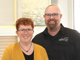 Raumausstattung Weist Ein lächelndes Paar, beide mit Brille. Die Frau trägt eine gelbe Jacke, der Mann ein schwarzes Hemd.