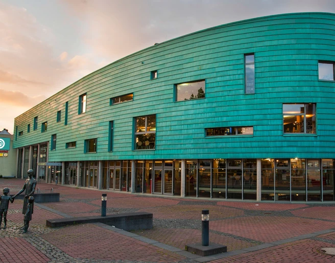 MEDIO Aussenansicht Daemmerung Futuristisches Gebäude mit grüner Fassade und Glasfront bei Sonnenuntergang auf einem Platz.</p>Futuristic building with green façade and glass front at sunset on a square.</p> <p