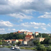 Blick auf die Altstadt von Pirna Stadtansicht mit Fluss im Vordergrund, historischen Gebäuden mit roten Dächern und grünem Turm, umgeben von Bäumen unter blauem Himmel mit Wolken.