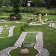 Minigolfbahnen am Fuß der Festung Königstein Minigolfplatz mit verschiedenen Bahnen und Hindernissen, umgeben von grüner Wiese und Bäumen; ruhige, einladende Atmosphäre.