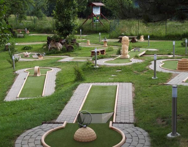 Minigolfbahnen am Fuß der Festung Königstein Minigolfplatz mit verschiedenen Bahnen und Hindernissen, umgeben von grüner Wiese und Bäumen; ruhige, einladende Atmosphäre.