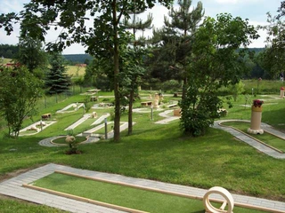 Minigolf in Königstein Eine Minigolfanlage mit mehreren Bahnen, umgeben von grünen Bäumen und Rasenflächen, unter einem leicht bewölkten Himmel.