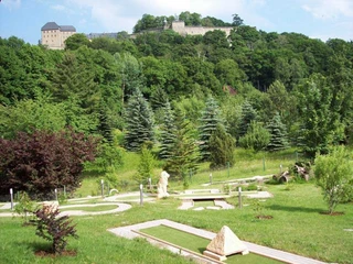 Minigolfen met uitzicht op de vesting Königstein Minigolfplatz in grüner Landschaft mit Bäumen und Sträuchern, im Hintergrund eine Burg auf einem bewaldeten Hügel.