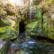 Amselfall Ein kleiner Wasserfall fließt über moosbedeckte Felsen in eine Höhle, umgeben von üppiger Vegetation und Steinmauern, in einer ruhigen, natürlichen Umgebung.