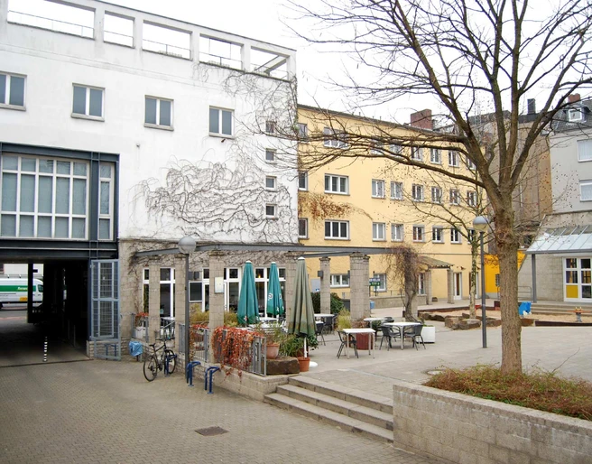 Ansicht Bürgerhaus Kalk aus dem hausinternen Hof Ein ruhiger städtischer Innenhof mit mehreren Gebäuden, Tischen und Stühlen im Freien.A quiet urban courtyard with several buildings, outdoor tables and chairs.