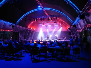 Open Air Bühne im Hof des BHK Open-Air-Bühne im Innenhof des BHK: Eine nächtliche Szene mit beleuchteter Bühne unter einem geschwungenen Zelt, umgeben von sitzendem Publikum.Open-air stage in the inner courtyard of the BHK: a nocturnal scene with an illuminated stage under a curved tent, surrounded by a seated audience.