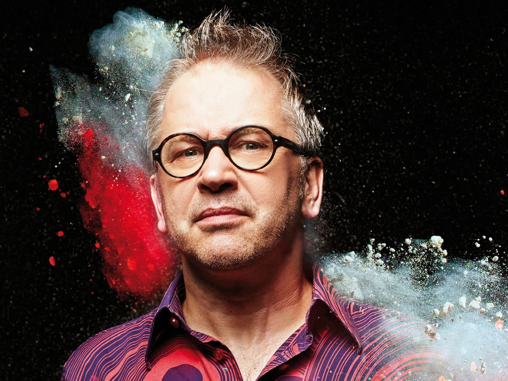 Robert Griess Porträt eines Mannes mit Brille vor schwarzem Hintergrund, umgeben von buntem Farbpulver.Portrait of a man with glasses in front of a black background, surrounded by colored powder.