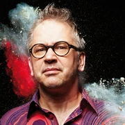 Robert Griess Porträt eines Mannes mit Brille vor schwarzem Hintergrund, umgeben von buntem Farbpulver.Portrait of a man with glasses in front of a black background, surrounded by colored powder.