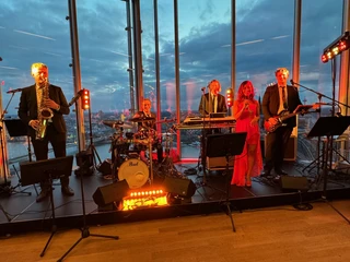 Confetti & Friends Eine sechsköpfige Band spielt vor einer gläsernen Skyline bei Sonnenuntergang.A six-piece band plays in front of a glass skyline at sunset.