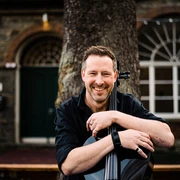 Simply Cello Mann lächelt, das Cello entspannend halten vor historischer Backsteingebäude-Fassade.Man smiling, holding the cello relaxing in front of a historic brick building façade.