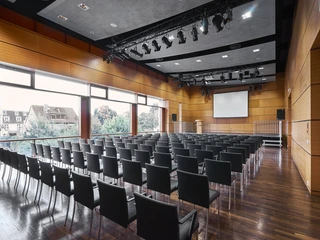 Kulturzentrum_kleiner_Saal_068.jpg Moderner Konferenzraum mit Holzwänden, leeren schwarzen Stühlen und einer großen Leinwand.</p>Modern conference room with wooden walls, empty black chairs and a large screen.</p> <p