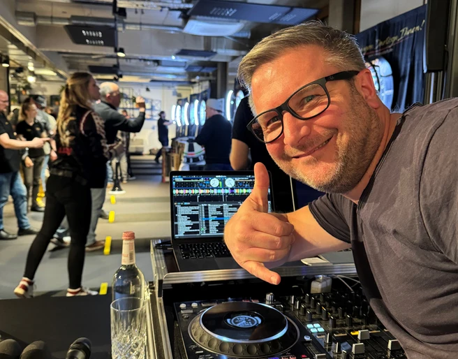 DJ Mr. Easy Ein DJ mit Brille vor einem Mischpult, das eine Veranstaltung mit Leuten im Hintergrund zeigt.</p>A DJ with glasses in front of a mixing desk showing an event with people in the background.</p> <p