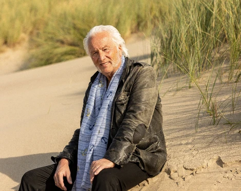 Hannes Schöner Alter Mann mit weißem Haar und Schal sitzt lächelnd im Sand vor Dünenlandschaft.Old man with white hair and scarf sits smiling in the sand in front of a dune landscape.