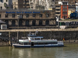 Kölntourist passenger boat service Das Bild zeigt ein großes weißes Riesenrad, das prominent in der Skyline von Köln am Rhend im Hintergrund histinufer thront. Im Vordergrund ist ein Ausflugsschiff auf dem Rhein zu sehen, währeorische Backsteingebäude das Stadtbild prägen.The picture shows a large white Ferris wheel, prominently enthroned in the skyline of Cologne on the banks of the Rhine in the background. An excursion boat on the Rhine can be seen in the foreground, while the cityscape is dominated by historic brick buildings.