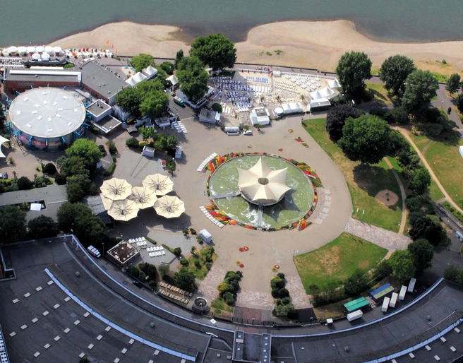 Tanzbrunnen Köln mit Rheinterrassen und km 689 Cologne Beach Club, Luftaufnahme Das Bild zeigt eine Veranstaltungslocation am Rhein mit einem sternförmigen Pavillon und umliegenden Grünflächen.The picture shows an event location on the Rhine with a star-shaped pavilion and surrounding green areas.