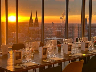 Firmenfeier_KölnSKY Köln, Domblick bei Sonnenuntergang durch Fenster, gedeckte Restauranttische vorne.Cologne, view of the cathedral at sunset through the window, covered restaurant tables in front.