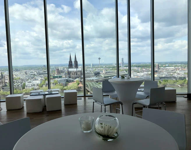Firmenfeier_KölnSKY Blick aus Panoramafenster auf die Kölner Skyline mit Dom, Fluss und Wolkenhimmel.</p>View from the panoramic window of the Cologne skyline with cathedral, river and cloudy sky.</p> <p