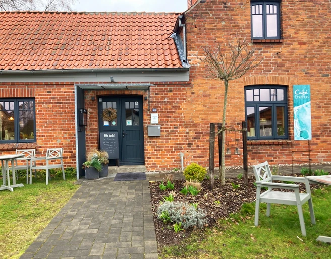 Cafe_EneHus03_032024_PetraRoemer.jpg Rotes Ziegelhaus mit Gehweg davor und Sitzecken links und rechts