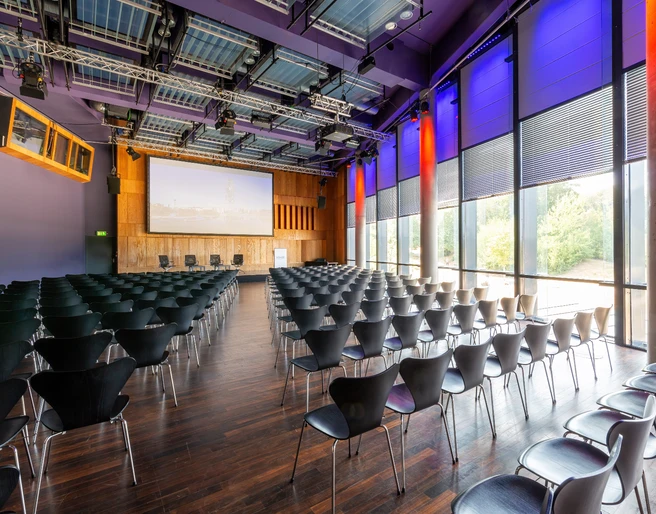 KOMED-Saal Moderner Konferenzraum mit dunklen Stühlen, großem Bildschirm und lila beleuchteten Wänden.</p>Modern conference room with dark chairs, large screen and purple-lit walls.</p> <p