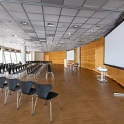 Raum 1-3  Ein moderner Konferenzraum mit leeren Stuhlreihen und zwei großen Bildschirmen.</p>A modern conference room with empty rows of chairs and two large screens.</p> <p