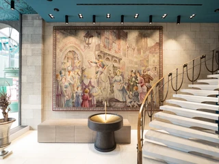 Dufthaus Interior 1 Wandteppich in historischem Gebäude, zeigt lebendige Straßenszene; elegante Treppe im Vordergrund.Tapestry in a historic building, showing a lively street scene; elegant staircase in the foreground.