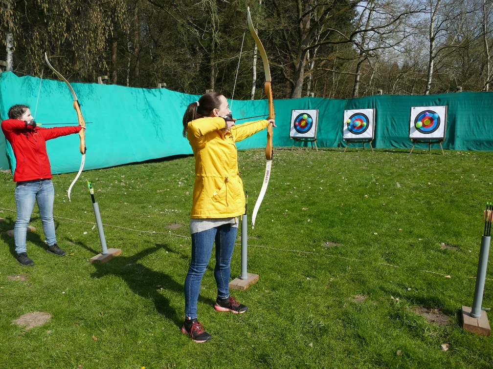 Bogenschießen.JPG Zwei junge Frauen stehen auf einer grünen Wiese und schießen mit dem Bogen auf Zielscheiben. Die Bögen sind gespannt und schussbereit.Two young women stand on a green meadow and shoot at targets with their bows. The bows are strung and ready to shoot.