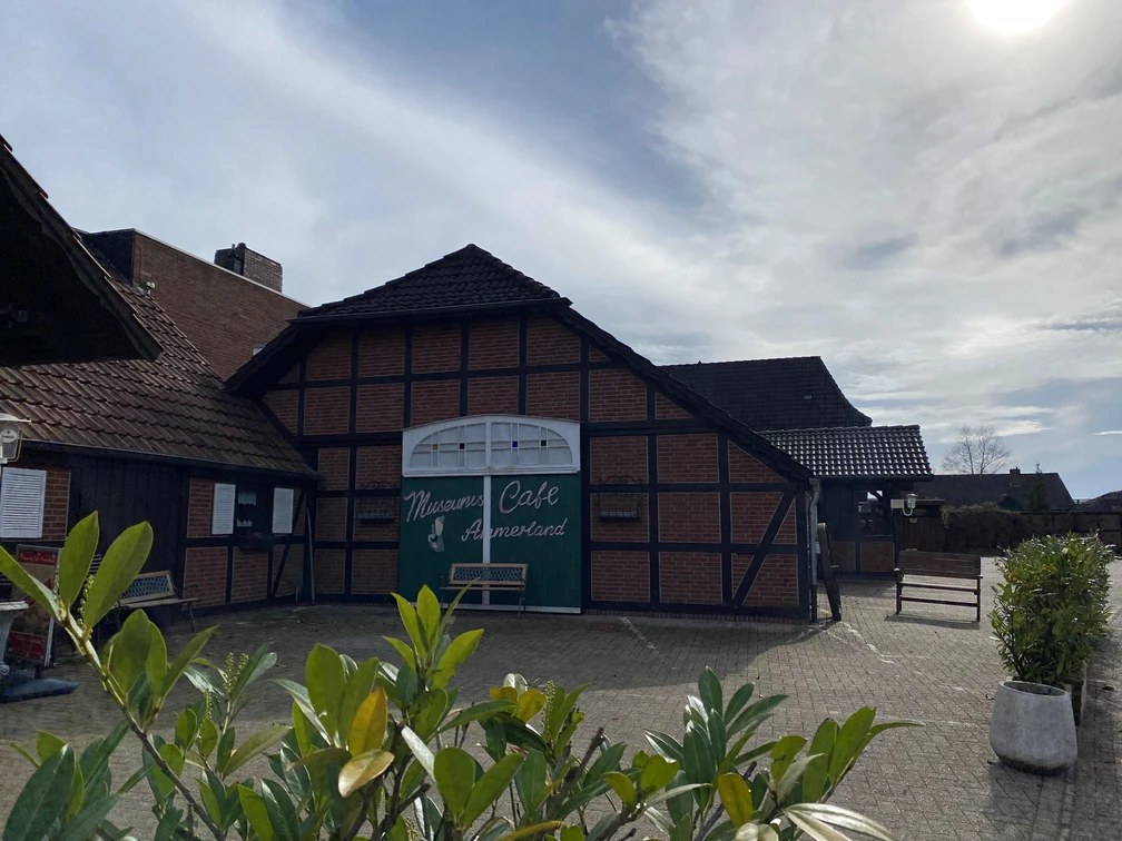 Bild 4.jpg Das Bild zeigt ein traditionelles Fachwerkgebäude mit der Aufschrift "Mühlen Café Ammerland". Der Vordergrund sind Pflanzen zu sehen, die dem Bild eine natürliche Note geben.