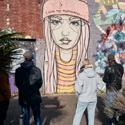 Ehrenfeld Street Art Tour Eine Gruppe von Menschen betrachtet ein großes Street-Art-Gemälde auf einer Backsteinmauer, das eine melancholische, junge Frau in Mütze zeigt. Im Vordergrund sind Pflanzen sichtbar.A group of people is looking at a large street art painting on a brick wall, depicting a melancholic young woman wearing a beanie. Plants are visible in the foreground.