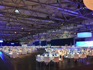 Abendveranstaltung Großer, elegant beleuchteter Bankettsaal mit weißgedeckten Tischen und glanzvollen Lichteffekten.</p>Large, elegantly lit banquet hall with white-clothed tables and glamorous lighting effects.</p> <p
