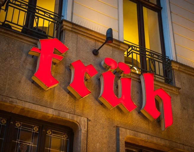 Brauhaus FRÜH am Dom Leuchtendes, rotes "Früh"-Schild an historischer Gebäudefassade bei warmem Abendlicht.</p>Bright red "Früh" sign on historic building façade in warm evening light</p>.
