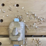 kasper-schnitzkopf-werkbank-schnitzbude-hohnstein-kasper-(c)-tvssw-madlen-rogge.jpeg Holzskulptur eines Gesichts in Arbeit auf einer Werkbank, umgeben von Holzspänen.