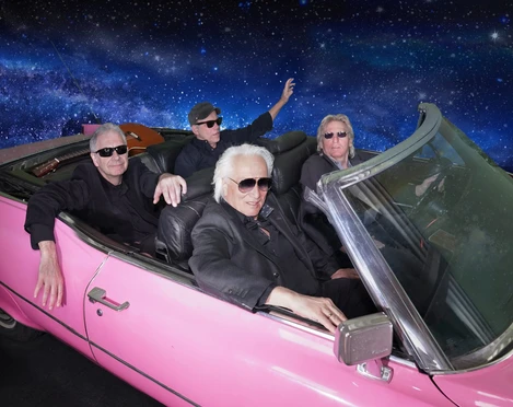 Hannes Schöner Band Vier ältere Männer in Sonnenbrillen fahren in einem pinken Cabrio durch eine Galaxie-Kulisse.Four older men in sunglasses drive through a galaxy backdrop in a pink convertible.