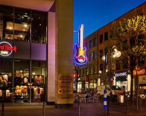 HRC Cologne_Exterior Außenansicht des Hard Rock Cafes mit beleuchteter Gitarre und umliegenden Geschäften in der Nacht.</p>Exterior view of the Hard Rock Cafe with illuminated guitar and surrounding stores at night.</p> <p