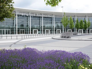 Congress-Centrum Koelnmesse Nord, Eingang Nordhallen Glasgebäude mit Eingangsbereich, flankiert von Bäumen und lila Blumen im Vordergrund.</p>Glass building with entrance area, flanked by trees and purple flowers in the foreground.</p> <p