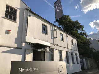 Hafen 12 Eingang eines weißen Gebäudes mit Mercedes-Benz-Logo und wehender Fahne bei Sonnenuntergang.</p>Entrance of a white building with Mercedes-Benz logo and waving flag at sunset.</p> <p