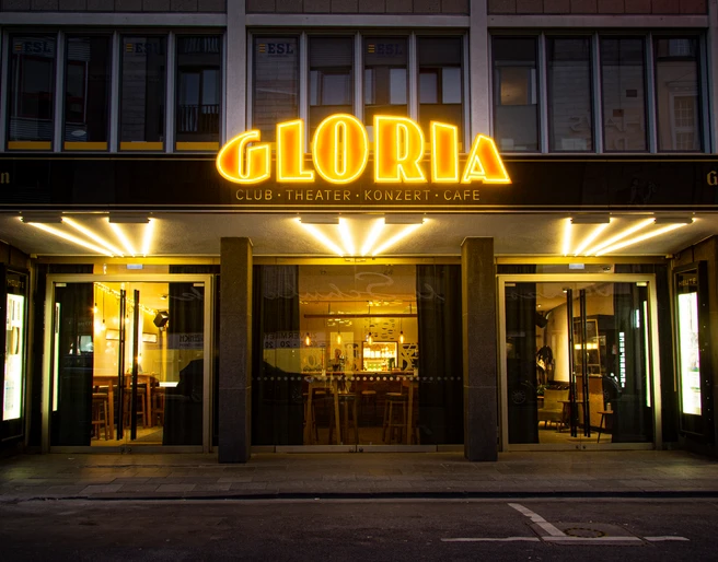GLORIA Außenansicht des beleuchteten Gloria Theaters mit auffälliger goldener Neonbeschriftung.</p>Exterior view of the illuminated Gloria Theater with eye-catching golden neon lettering.</p> <p