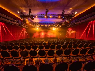 A7R00450.jpg Beleuchteter Theatersaal mit leeren Stuhlreihen und rotem Samthintergrund; Bühne im Licht.</p>Illuminated theater hall with empty rows of chairs and red velvet background; stage in the light.</p> <p