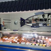 Allräucherei Prahm-Theke-Apen.jpg Ein Verkäufer in weißem Kittel grüßt freundlich hinter einer gut bestückten Fischtheke.