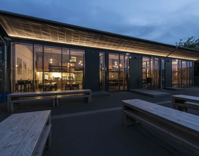 DEINspeisesalon Eventlocation Moderne Glasfassade eines Gebäudes in der Dämmerung, beleuchtet mit warmen Lichtern im Inneren.</p>Modern glass façade of a building at dusk, illuminated with warm lights inside.</p> <p