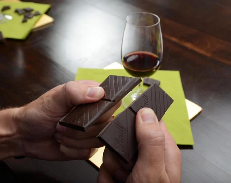 Verkostung Wein & Schokolade 2 Person bricht dunkle Schokolade, daneben ein Glas Rotwein auf einem grünen Untersetzer.</p>Person breaks dark chocolate, next to it a glass of red wine on a green coaster.</p> <p