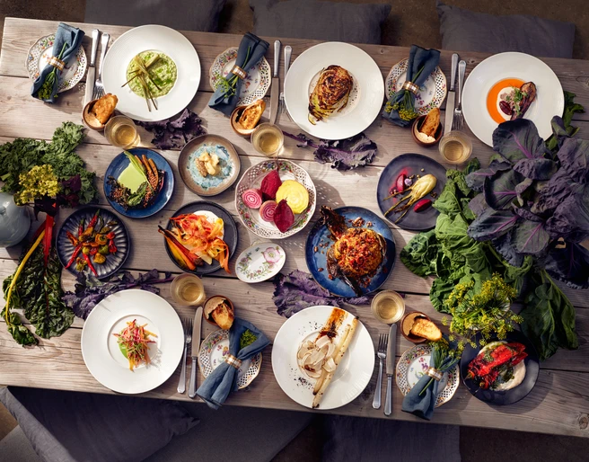CATERING NEU DENKEN Farbenfrohe, abwechslungsreich angerichtete Speisen auf einem hölzernen Tisch, dekoriert mit Pflanzen.</p>Colorful, varied dishes served on a wooden table, decorated with plants.</p> <p
