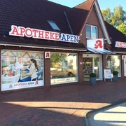 Apotheke-Apen_2023.jpg Das Bild zeigt die Frontansicht der Apotheke Apen mit einem markanten roten Schriftzug auf dem Dach. Die Fassade ist aus roten Backsteinen und verfügt über große Fenster, die Einblicke ins Innere gewähren. Vor der Apotheke befindet sich ein breiter Gehweg mit Pflastersteinen, begleitet von grünen Pflanzen in Töpfen. Die Apotheke strahlt eine einladende Atmosphäre aus.