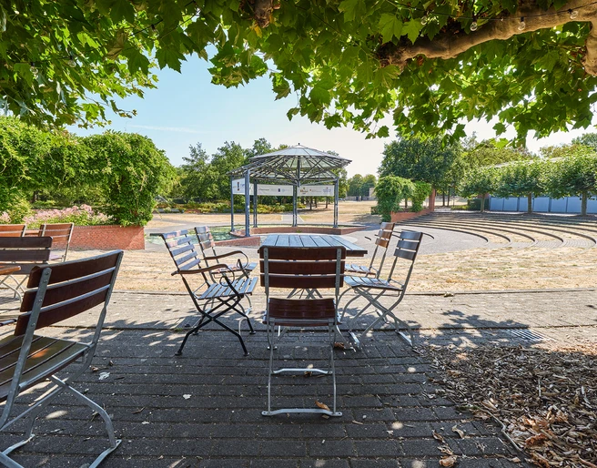 Ringlokschuppen Ruhr, Mülheim an der Ruhr <p>Ein Park mit leeren, hölzernen Bistrotischen und Stühlen unter grünen Bäumen, nahe einer Freilichtbühne.</p>
