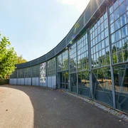 Ringlokschuppen Ruhr, Mülheim an der Ruhr Halbrundes Gebäude mit großflächigen Glasfassaden, Bäume und Sonnenstrahlen im Hintergrund.