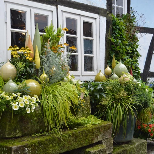 Üppig bepflanzte Steinstufen vor einem Fachwerkhaus mit dekorativer Keramik im Vordergrund.