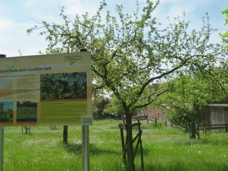 Hinweisschild zur Natur auf einer grünen Wiese mit Apfelbaum und einem rustikalen Gebäude im Hintergrund.