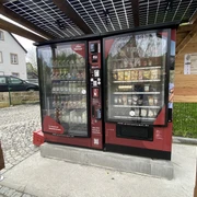Proviantomat Standort in Schöna Roter Verkaufsautomat unter Solardach, gefüllt mit Getränken und Snacks, steht auf gepflastertem Platz neben einem Haus und Holzwand.