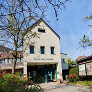 Haus des Gastes in Hinterhermsdorf Ein helles Gebäude mit der Aufschrift "Haus des Gastes", umgeben von blühenden Rosen und Bäumen, unter einem klaren blauen Himmel.