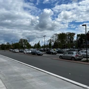 Parkplätze am Dockgelände.jpg Das Bild zeigt einen Parkplatz am Dockgelände an einem wolkigen Tag. Zahlreiche Autos sind auf dem asphaltierten Parkplatz geparkt, umgeben von ordentlich angelegten Gehwegen und Straßenlaternen. Im Hintergrund sind vereinzelte Bäume und Gebäude zu sehen, die eine urbane Umgebung andeuten.